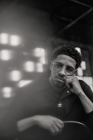 Monochrome portrait of adult male with glasses indoors. Man poses with curly hair, wearing black polo shirt, resting head on hand.