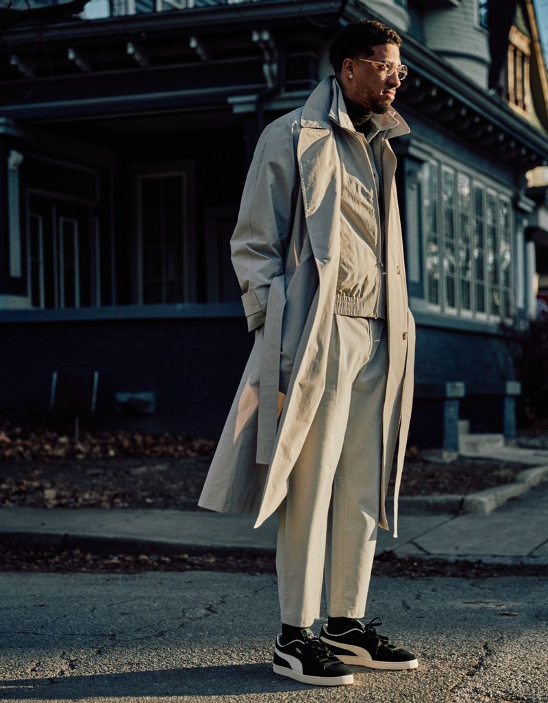 An adult male in a tan trench coat and pants standing on a city street in front of a dark, two-story home on a sunny day.