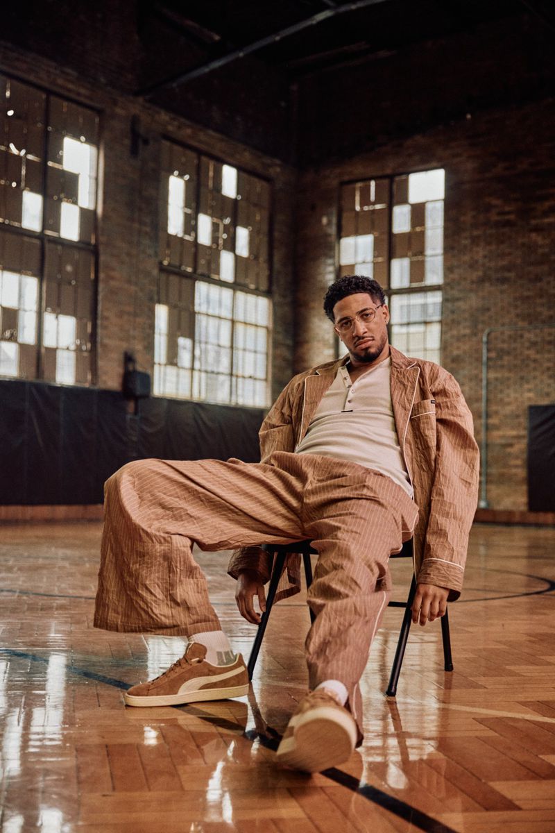 Man sitting in vintage gymnasium wearing matching tan separates with Puma sneakers, lit by large windows casting soft light.
