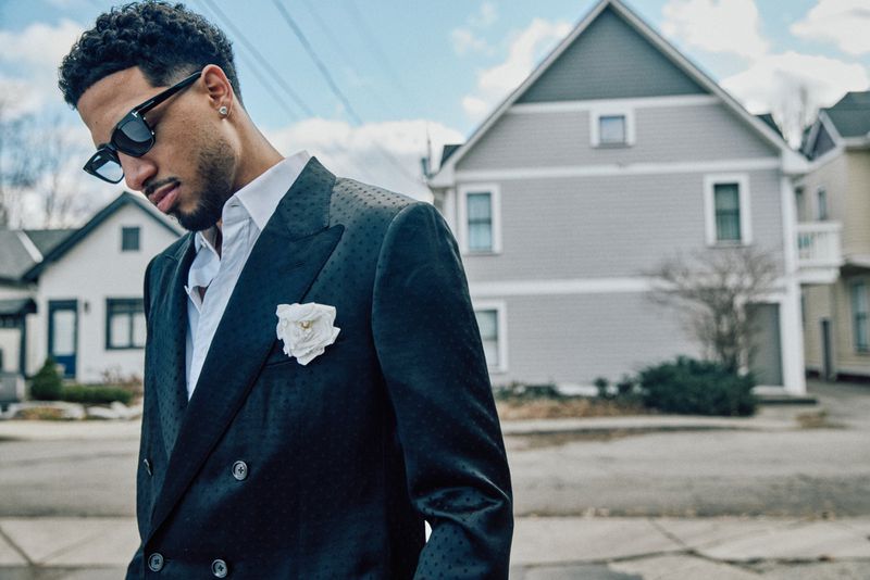 Stylish man in a dark suit, white shirt, and sunglasses poses on a city street with houses in the background under a bright sky.