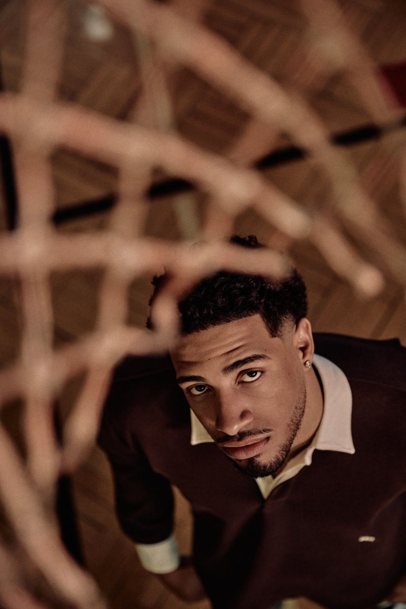Low-angle portrait of a young adult man looking up with a basketball hoop and net visible in the foreground and background.