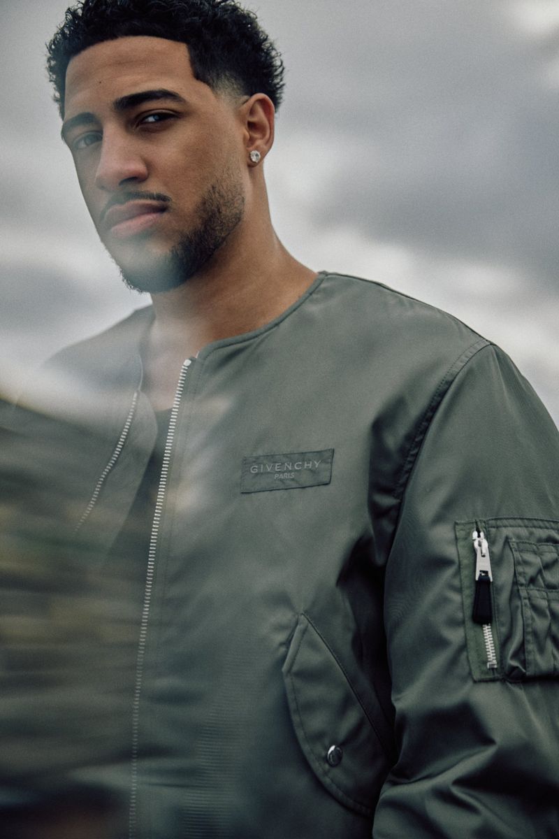 Close-up portrait of a young adult male wearing a green bomber jacket with a logo, set against an overcast sky, casual fashion.