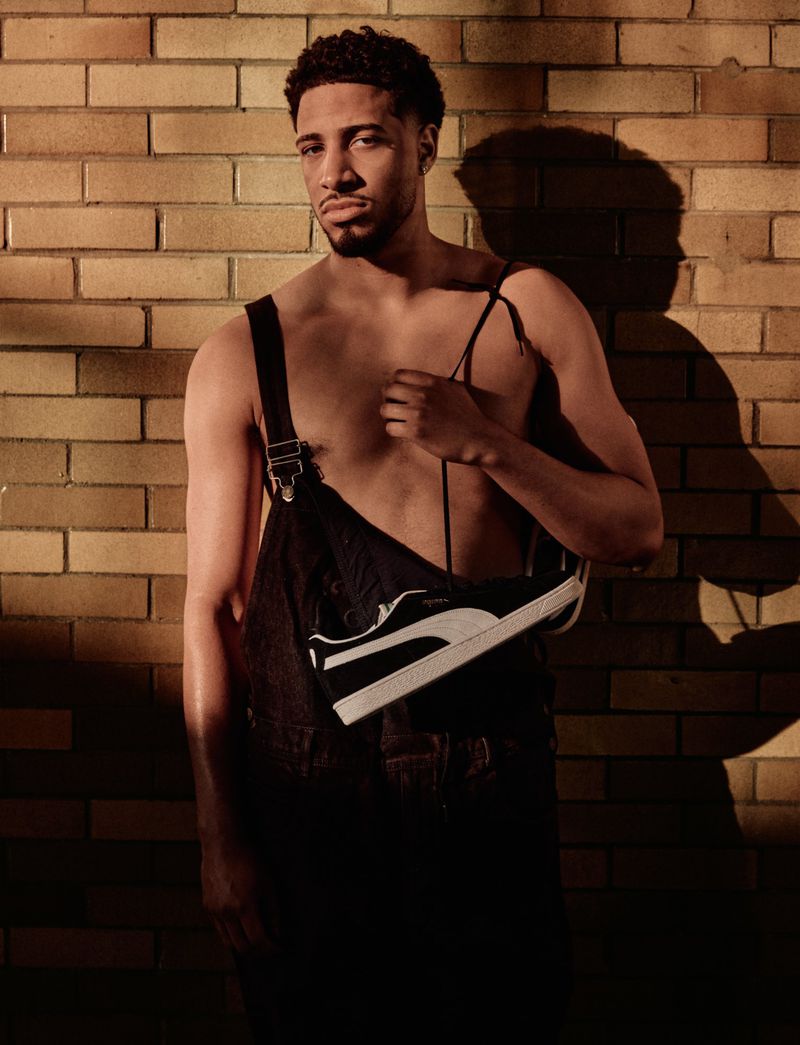 An adult male poses in front of a brick wall wearing black overalls and holding a black and white sneaker in a photo studio.