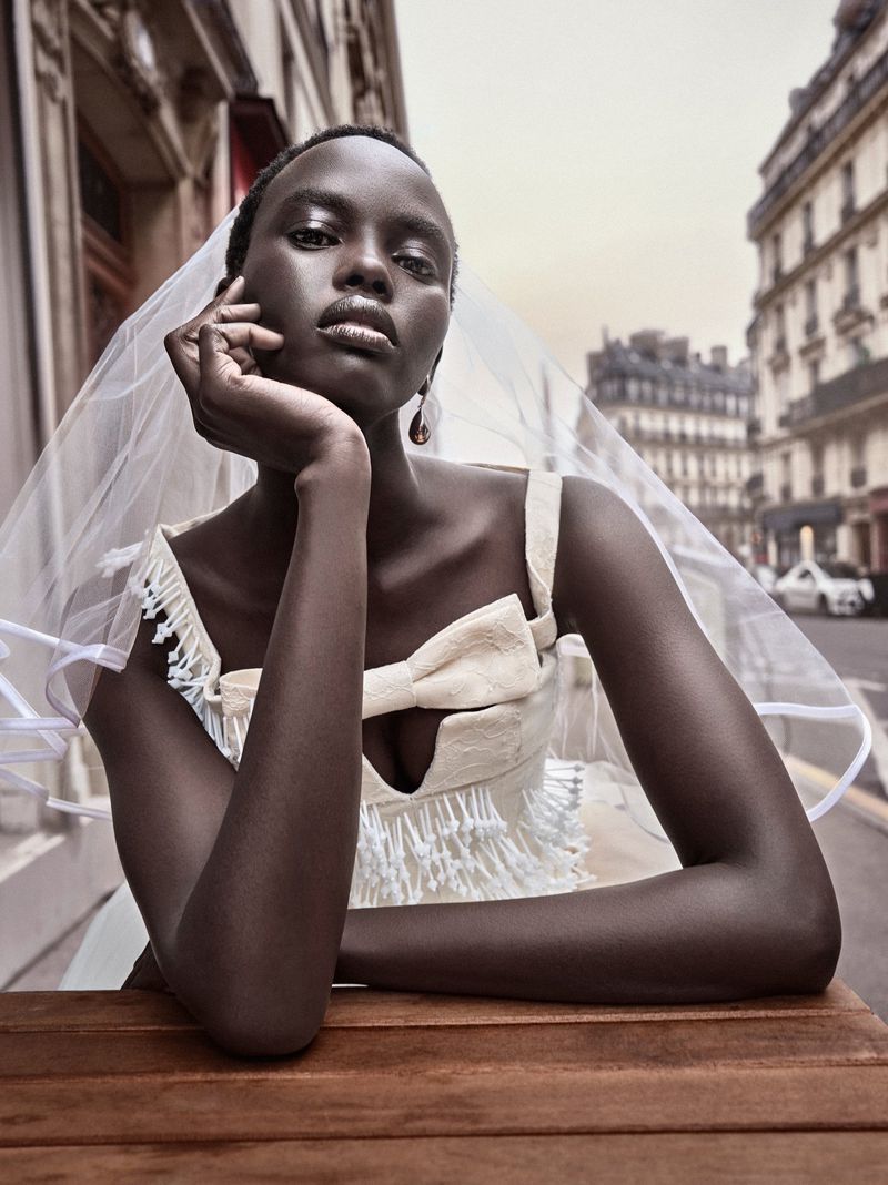 Elegant dark-skinned woman in white dress with bow, posing at a table in a European city with buildings in the background and soft light.