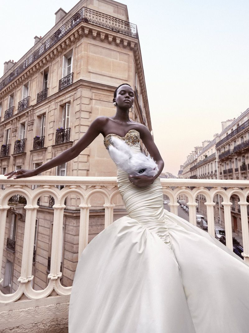 Fashion shot of an African American woman in a wedding dress holding a white rabbit in front of a Parisian building on a balcony.