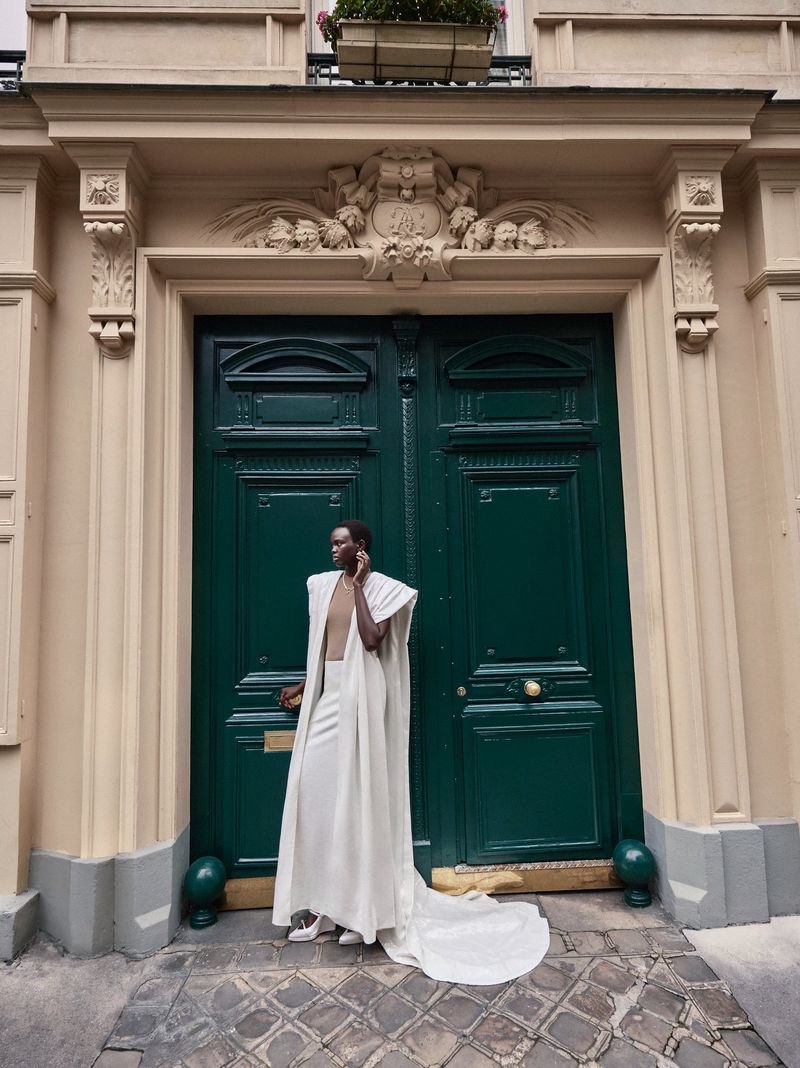 Person wearing cream-colored formalwear poses in front of a green door, with an off-white building exterior in an urban setting.