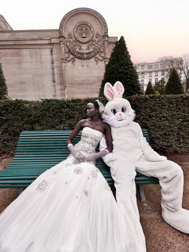 Fashion editorial featuring an African woman in wedding dress sitting next to a human in a rabbit costume on a green bench.