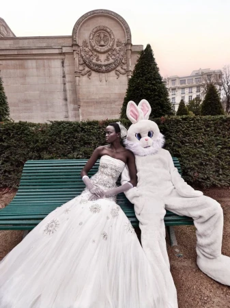 Fashion editorial featuring an African woman in wedding dress sitting next to a human in a rabbit costume on a green bench.