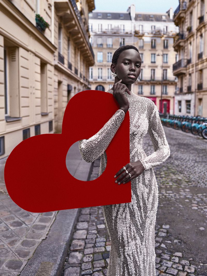 Elegant woman in sequined dress posing with large red heart prop in Paris street, showcasing fashion and urban scene with classic architecture.