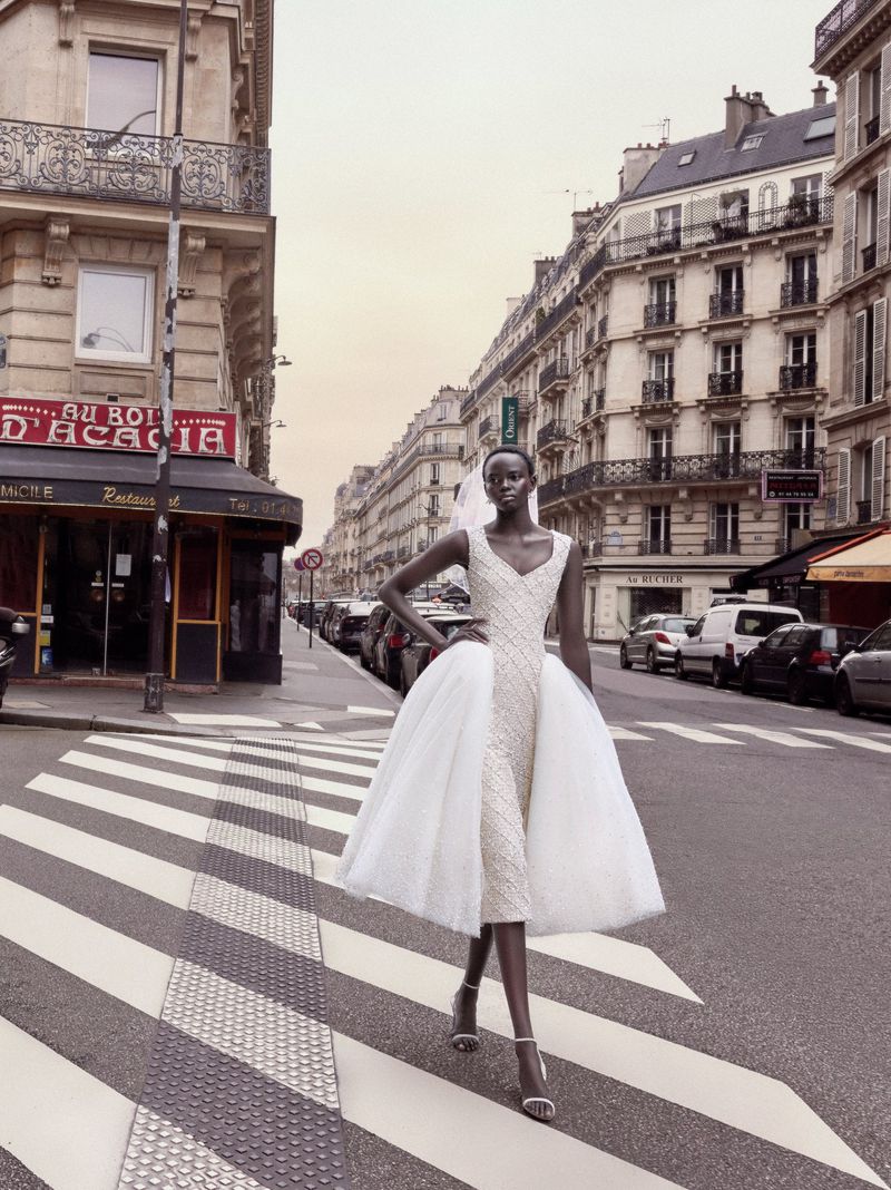 Woman in white dress walks on zebra crossing in urban setting, surrounded by buildings, parked cars, and street signs; outdoor fashion.