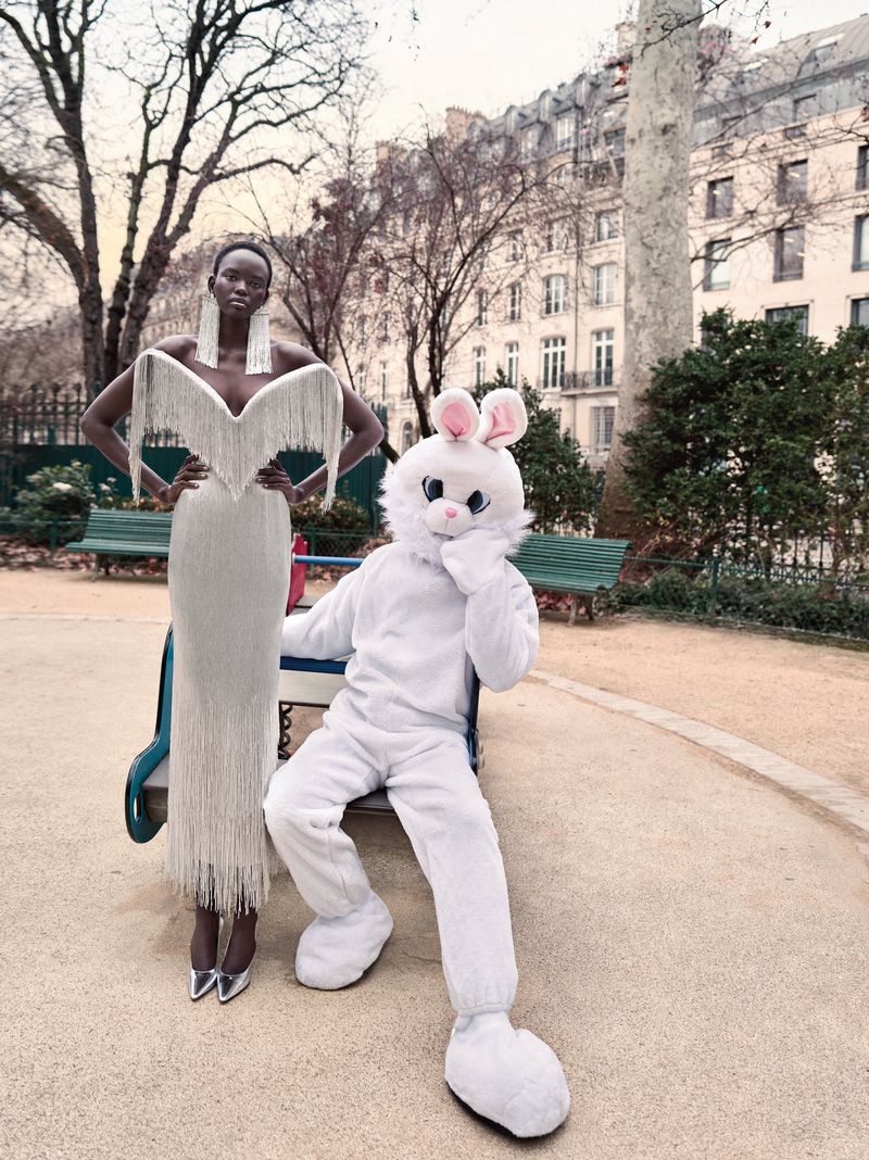 Fashion photo of an African-American model in a white gown posing with a person in a white bunny costume in a park.