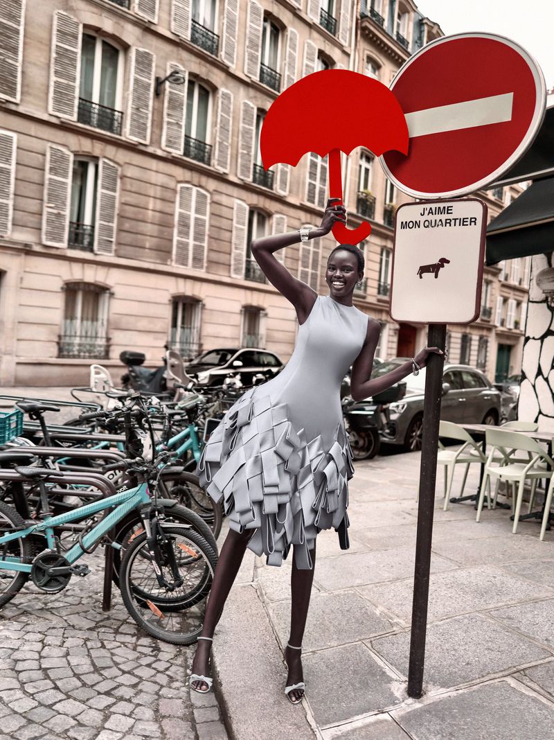Woman in gray dress poses with red umbrella sign on Paris street surrounded by bicycles and architecture in a fashion editorial.