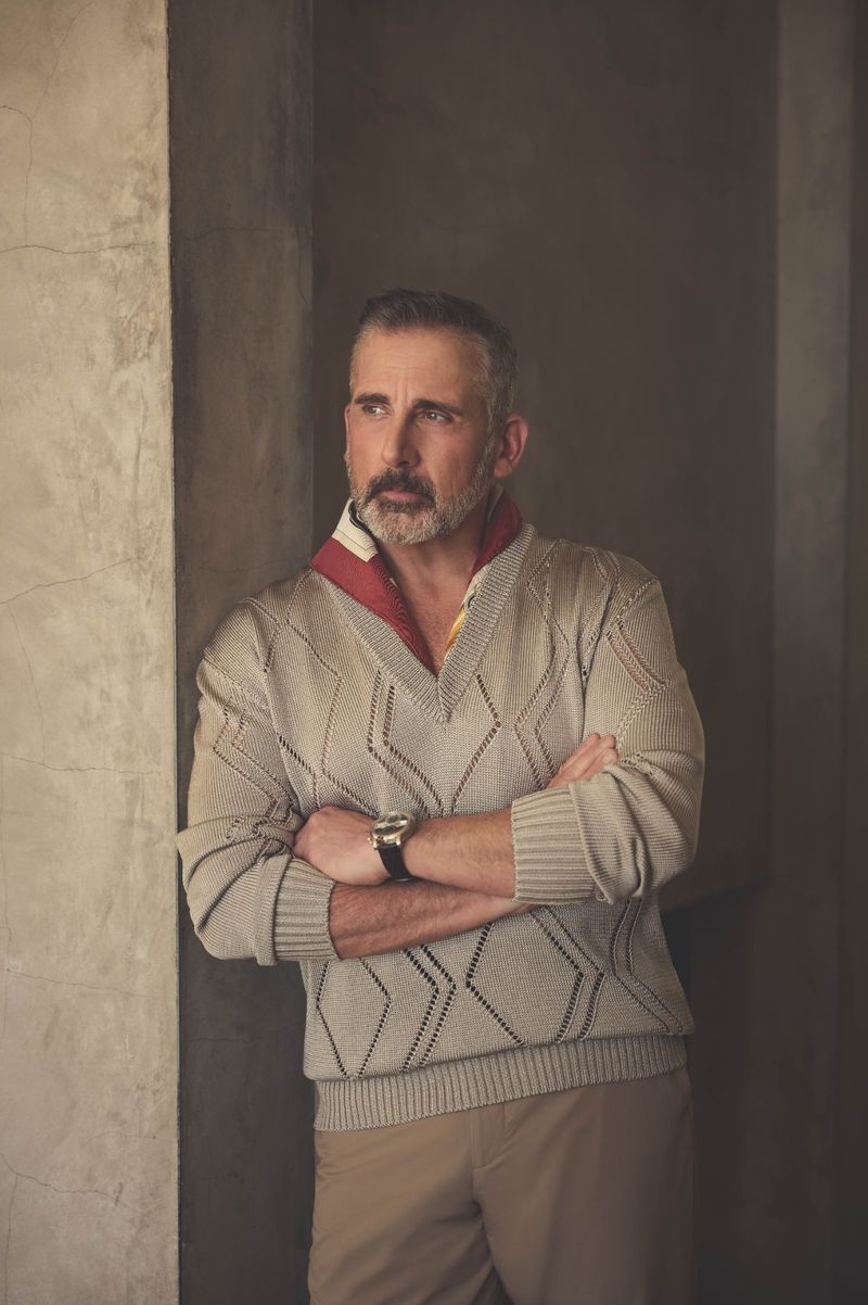 Adult man wearing a v-neck sweater stands indoors with his arms crossed, looking to the left, with a concrete wall in the background.