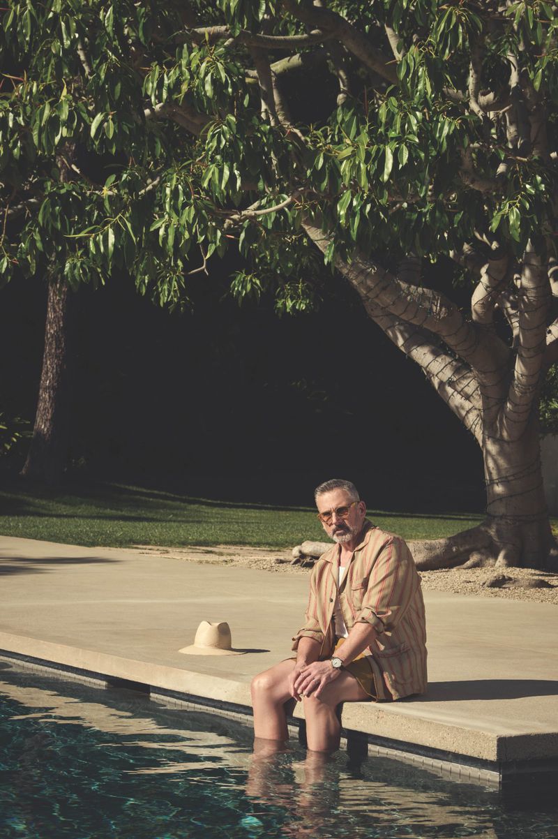 Mature man sits by a pool with his feet in the water on a sunny day, near a large tree, wearing casual clothing and sunglasses.