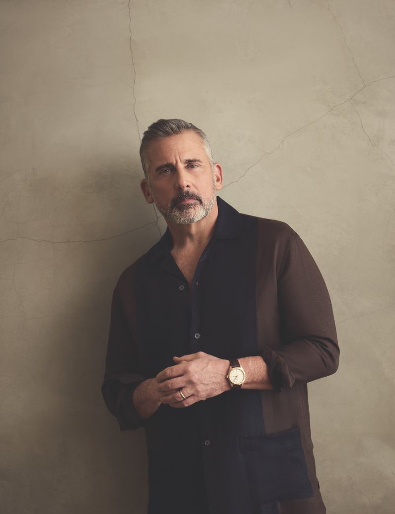 Portrait of a middle-aged man with graying hair and a beard, wearing a dark collared shirt and a gold watch against a textured wall.