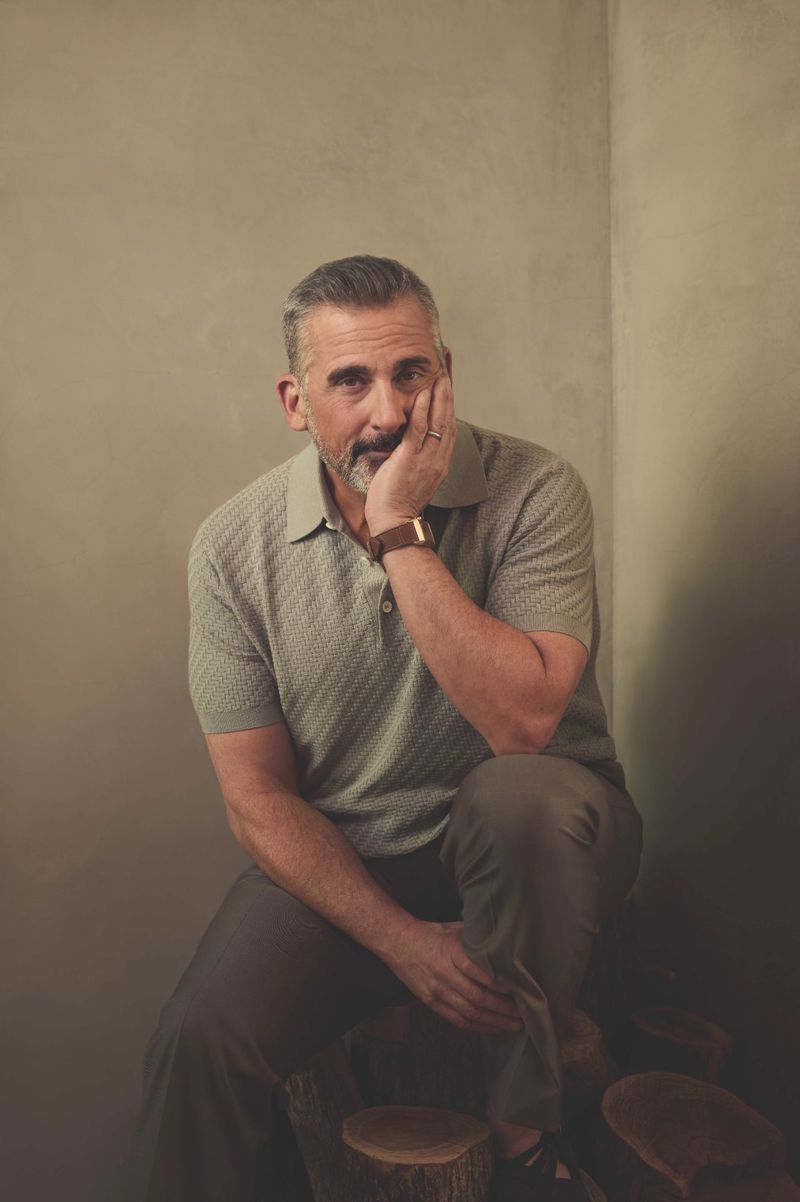 Portrait of a mature man with gray hair, wearing a sage green polo shirt, seated with his hand to his face in a neutral indoor setting.