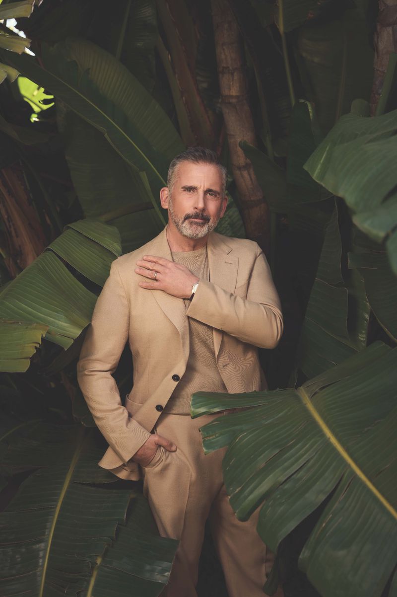 Stylish man in a tan suit poses amid vibrant tropical foliage, creating a portrait perfect for fashion and celebrity lifestyle content.