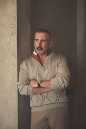 Steve Carell standing with arms crossed wearing a patterned beige knit sweater and a watch against a dark textured wall.