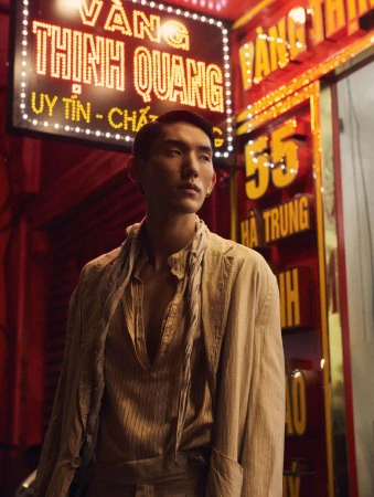 Asian man in a vintage linen outfit posing in front of a neon sign at night in an urban environment with red walls and dim lighting.