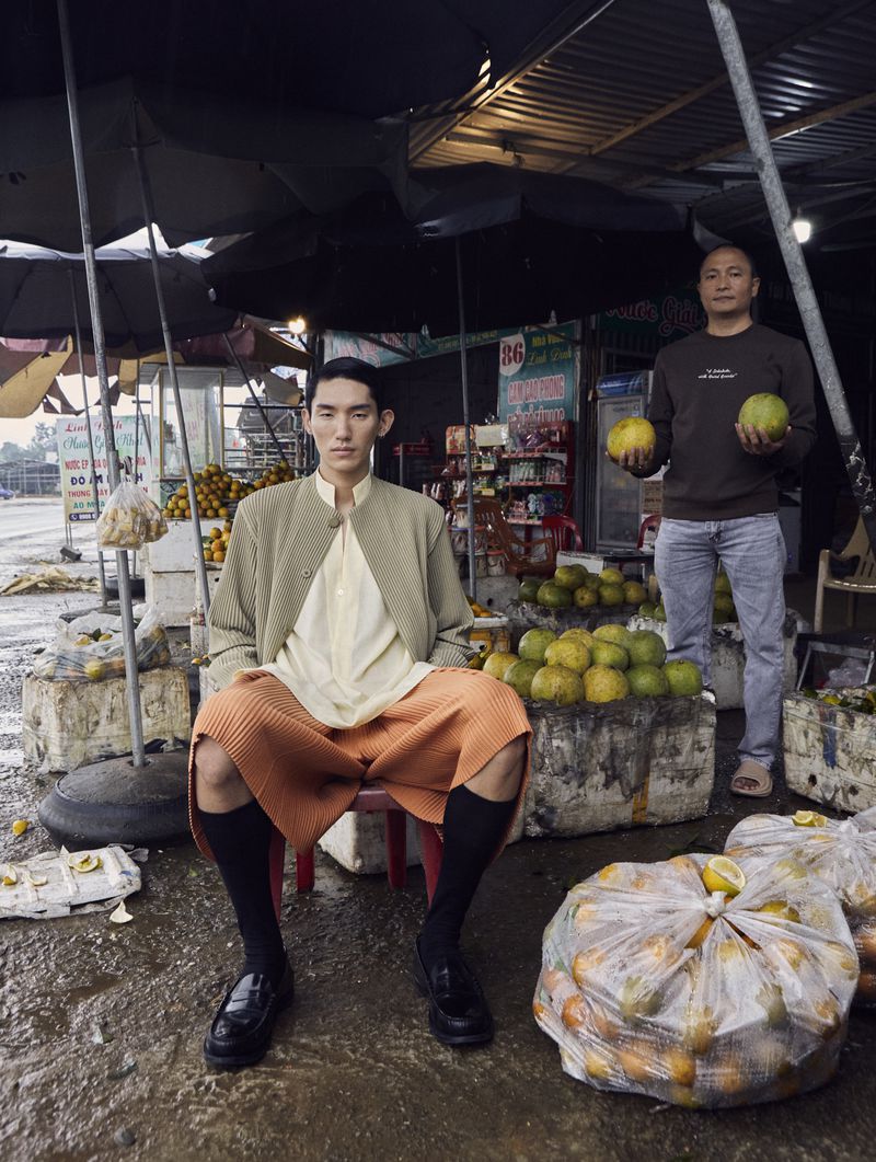 Asian male model seated at an outdoor market surrounded by fruit with a man holding a green fruit standing in the background.