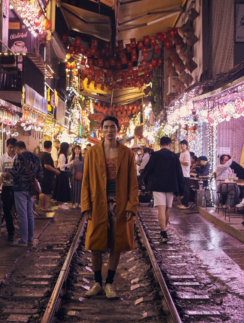 Adult male in orange coat poses on railroad tracks at night, surrounded by a crowd of people and festive red and white lights.