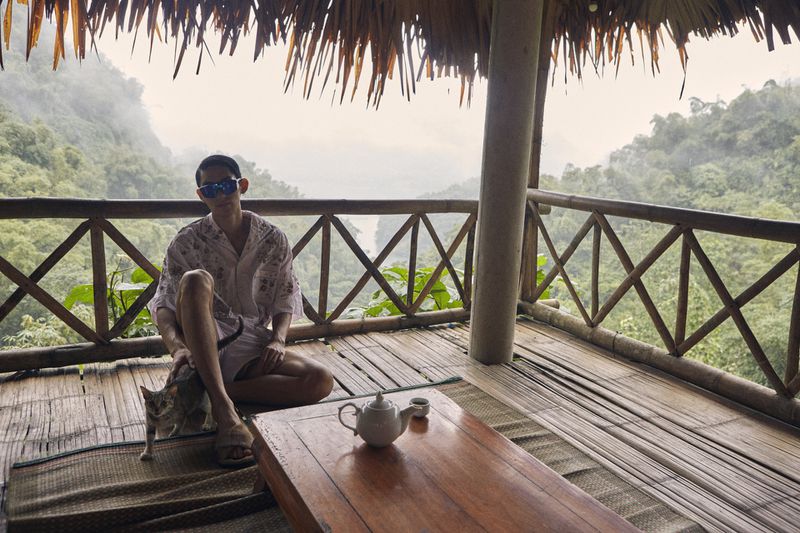 Person relaxing on a bamboo porch with a cat, a tea set, and a lush mountain view in the background on an overcast day.