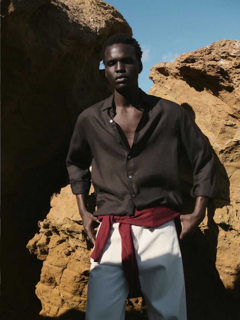 Model standing outdoors against rocky terrain wearing a dark brown shirt and white trousers with a red sweater tied around the waist.