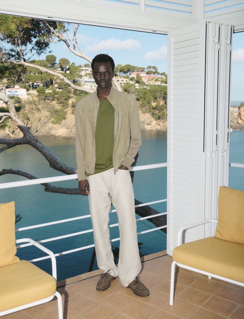 Adult male model poses on a white-railed porch, dressed in casual tan jacket and pants, overlooking the ocean and a green-treed coastline.