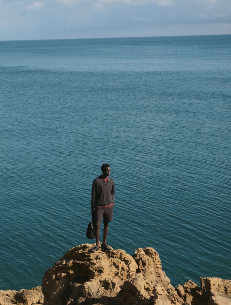 A man standing on a rocky outcrop overlooking a wide blue ocean under an open sky