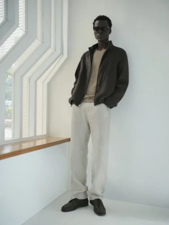 Dark-skinned adult male in sunglasses, brown jacket, light trousers, and black shoes, standing indoors with window lighting.