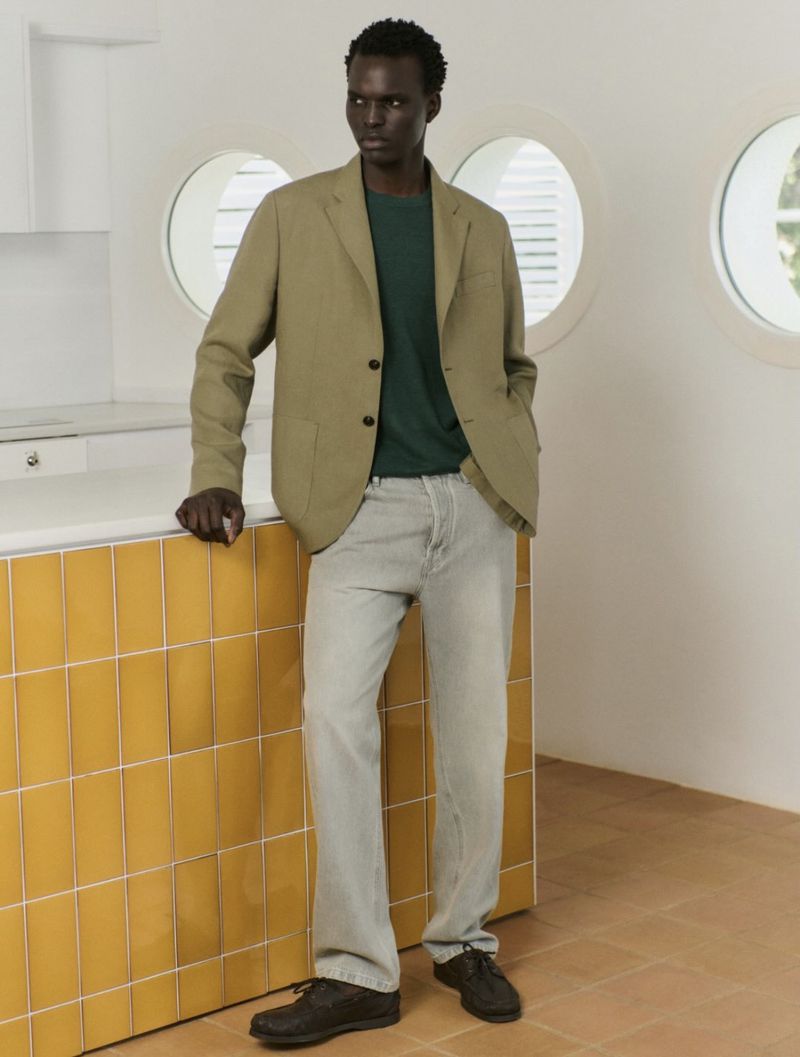 A dark-skinned man poses indoors wearing a tan blazer, green t-shirt, and grey jeans, leaning against a tiled counter with a minimalist background.