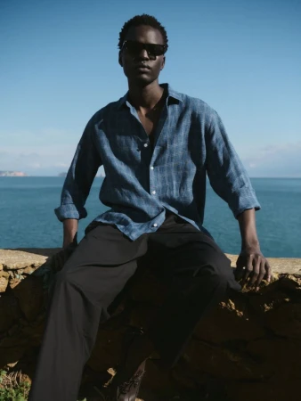 A man in sunglasses and a casual blue shirt sits on a stone ledge with ocean view in the background, casual men's fashion editorial.