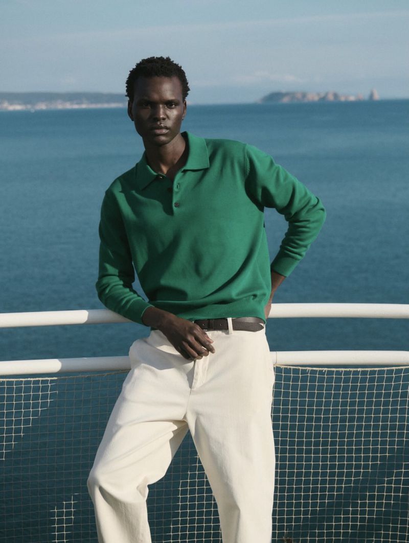 Dark-skinned man in green polo shirt and cream pants poses near a railing, with the ocean and distant hills visible in the background.