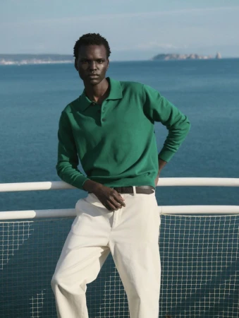 Dark-skinned man in green polo shirt and cream pants poses near a railing, with the ocean and distant hills visible in the background.