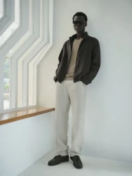 Black man wearing dark jacket and light trousers leaning against a white studio wall near a large window