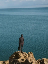 A man standing on a rocky outcrop overlooking a wide blue ocean under an open sky