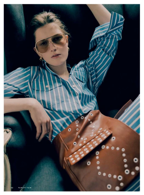A woman wearing a striped shirt and aviator sunglasses sits with a brown leather bag in an indoor setting, illuminated by natural light.