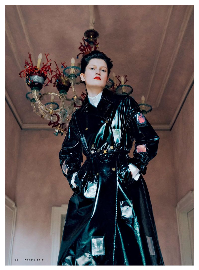 Fashion editorial image featuring a woman with short dark hair in a shiny black trench coat posing in front of an ornate chandelier.