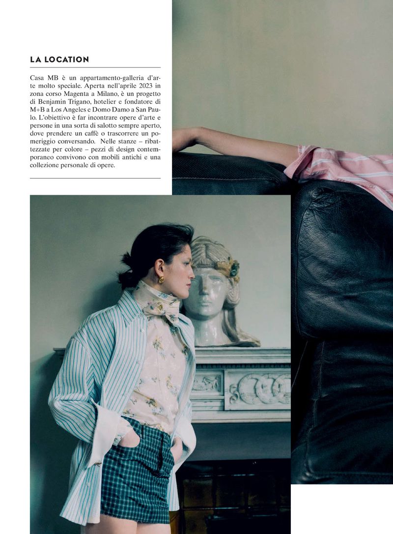 Editorial fashion shot featuring a stylish adult female in a striped jacket and plaid skirt posing indoors near a bust sculpture and couch.