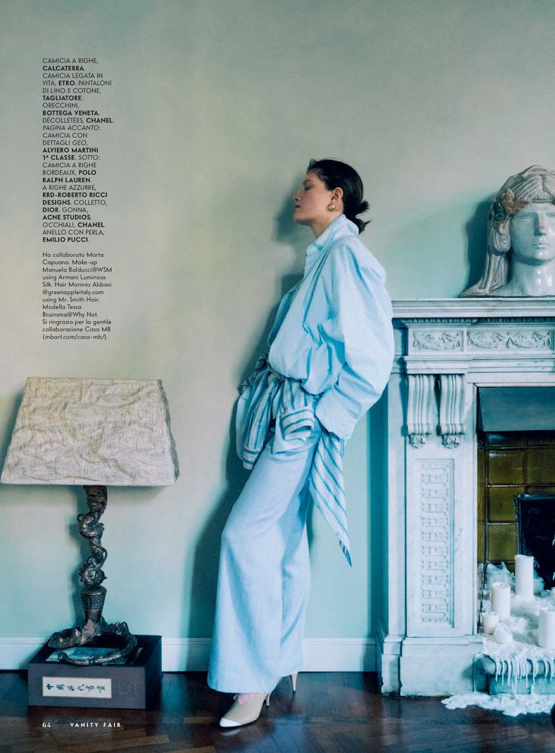 A woman in a light blue outfit poses in an interior with vintage decor, next to a lamp and fireplace with bust sculpture.