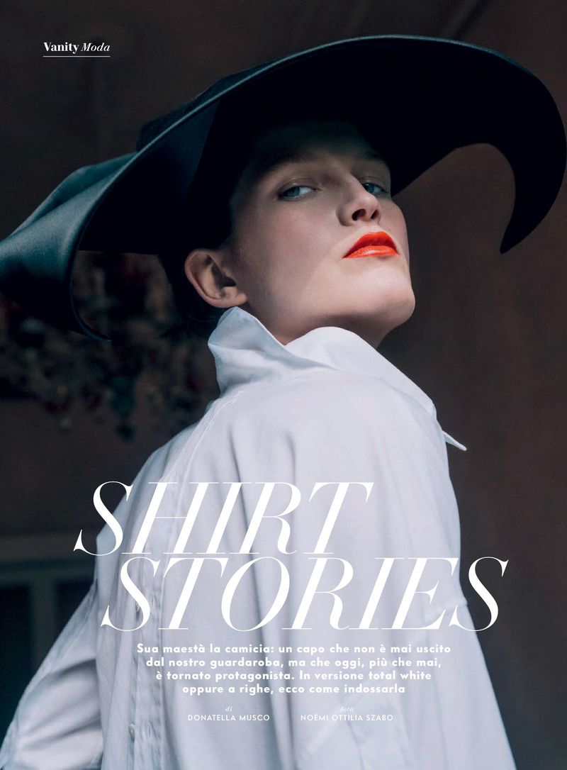Editorial fashion shot of a woman wearing a wide-brim hat and white collared shirt, accented by vivid orange lipstick and a low angle.