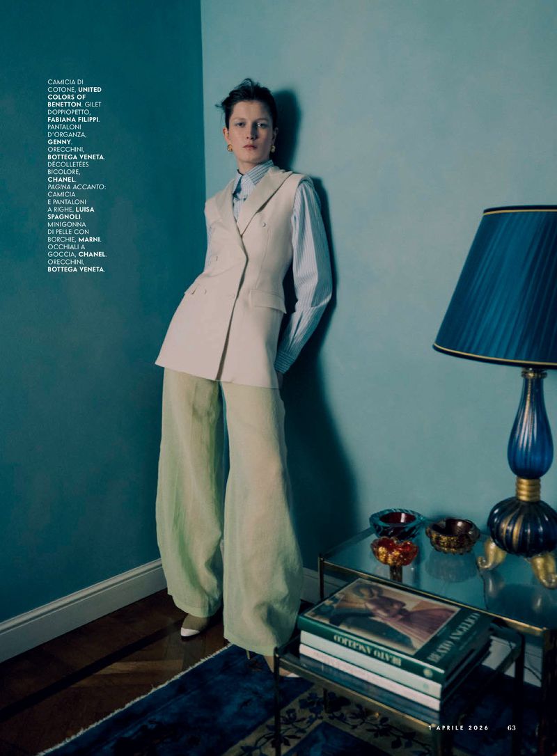 A woman poses indoors wearing a sleeveless beige blazer, light blue shirt, and pale green pants, styled with neutral makeup and a serious expression.