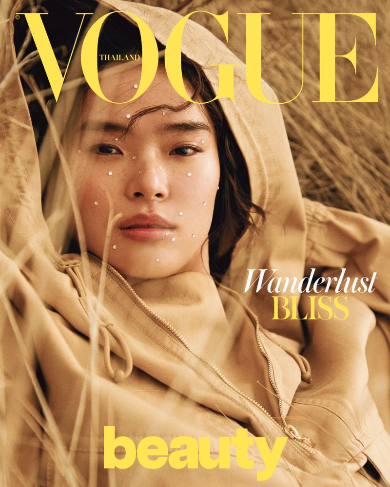 Vogue Thailand magazine cover featuring an Asian female model in a beige hooded jacket, adorned with crystals, lying in tall grass.