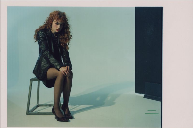 A redhead woman sits on a metal stool in a studio, wearing a black leather jacket, dress, sheer stockings, and patterned heels against a blue-white background.