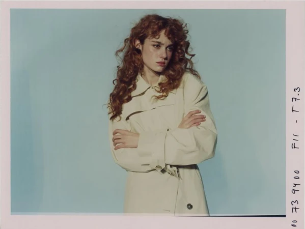 Portrait of a young adult woman with red curly hair wearing a beige trench coat, arms crossed, standing against a light blue background.