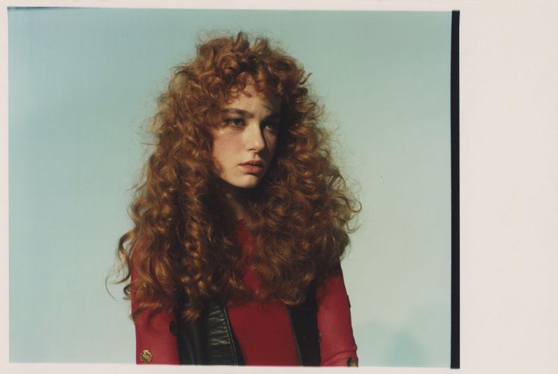 Portrait of a young woman with voluminous curly red hair wearing a red top and dark vest against a light blue background.