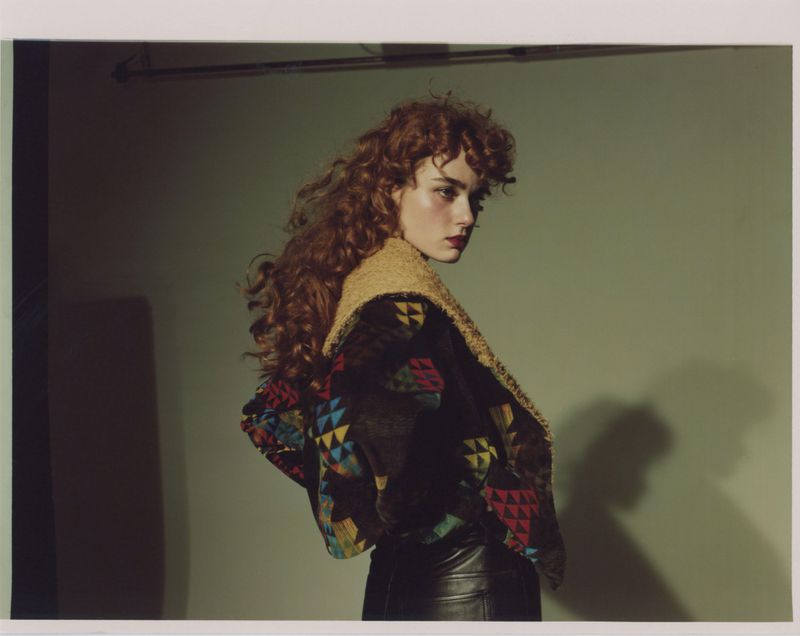 A woman with auburn curly hair in a patterned jacket looks over her shoulder against a neutral backdrop in a fashion editorial-style photo.
