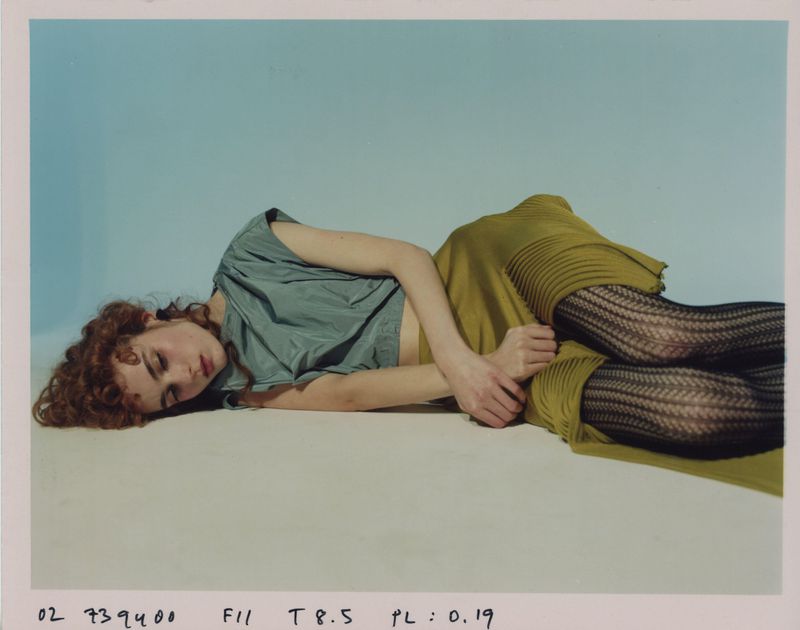 Woman with red hair poses on floor in gray top, chartreuse pleated skirt, and patterned black hosiery, against a pale blue background.