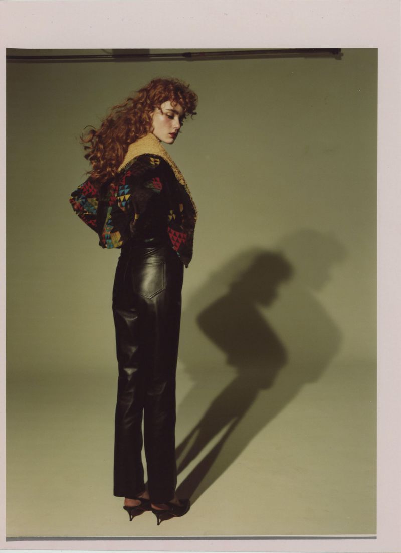 Fashion portrait of a woman with red curly hair wearing a black, embroidered jacket and leather pants, posing against a green backdrop.