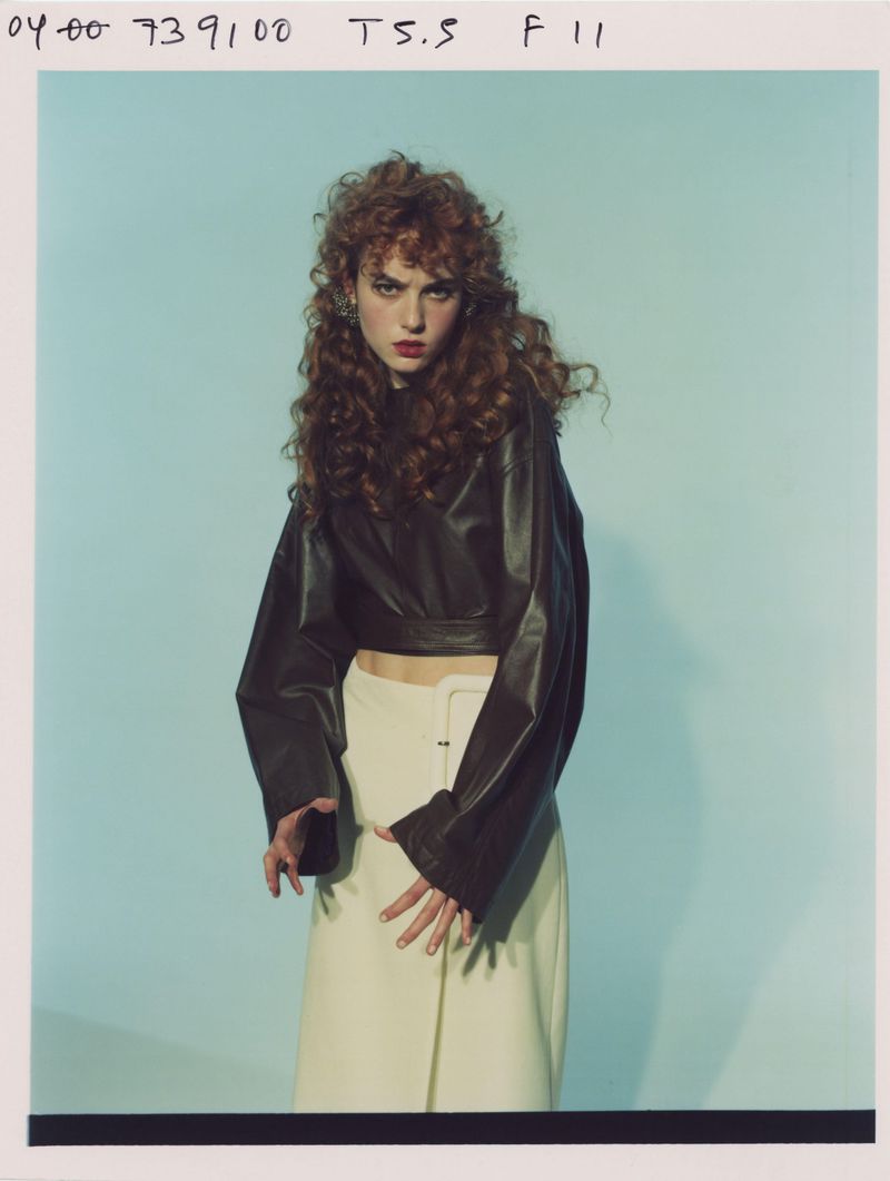 Model with long curly red hair wearing a dark leather top and white skirt against a blue studio backdrop.